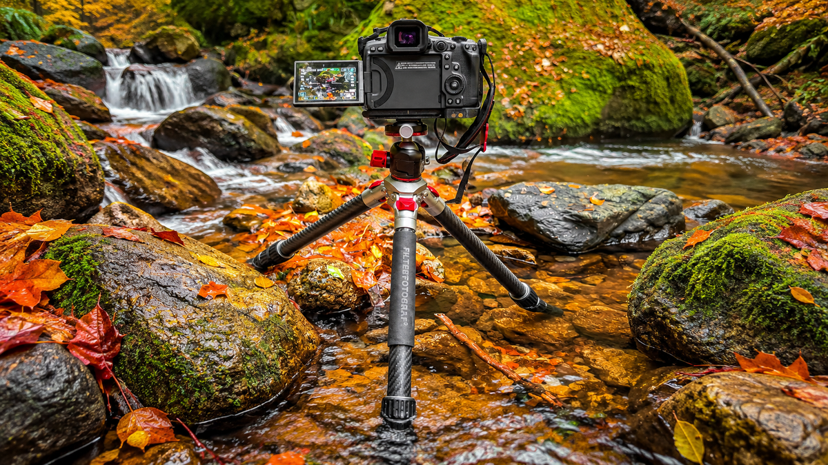FILTERFOTOGRAF FF1300 Carbon-Reisestativ mit montierter Kamera in einem flachen Waldbach, umgeben von moosbewachsenen Felsen und buntem Herbstlaub.