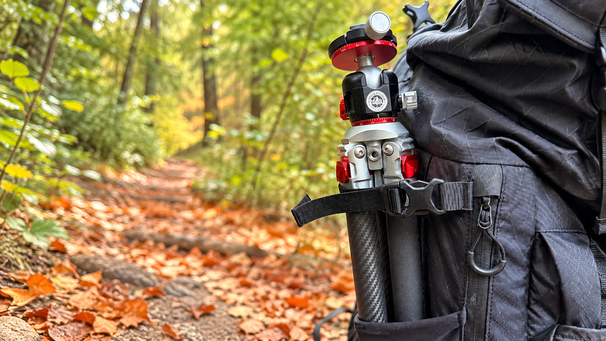 FILTERFOTOGRAF FF1300 Carbon-Reisestativ mit kompaktem Packmaß von 47 cm, platzsparend in der Seitentasche eines Kamerarucksacks verstaut, auf einem Waldweg.